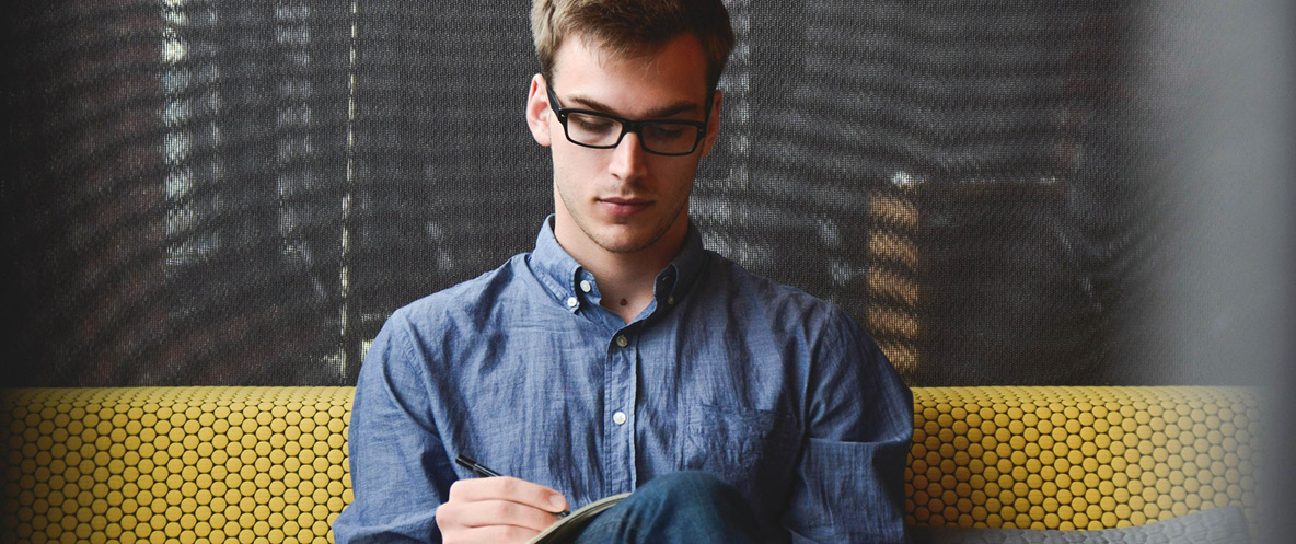 Young man with glasses on a couch writing in a notebook