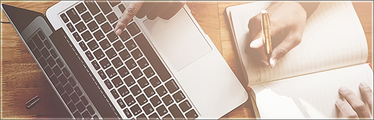 Laptop and a notebook with a hand pointing at the laptop and hands beginning to write in the notebook